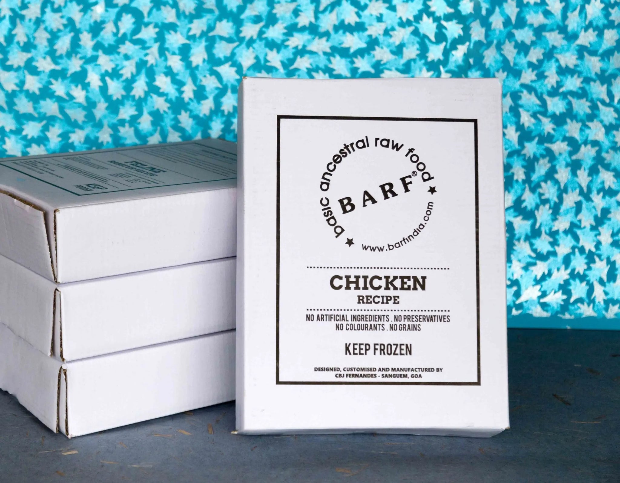 A box of www.barfindia.com's Raw Dog Food - Cage-Free Chicken Recipe for Complete Nutrition is in front of three stacked white boxes with a blue leaf-patterned background. This premium raw food contains no artificial ingredients and says Keep Frozen.