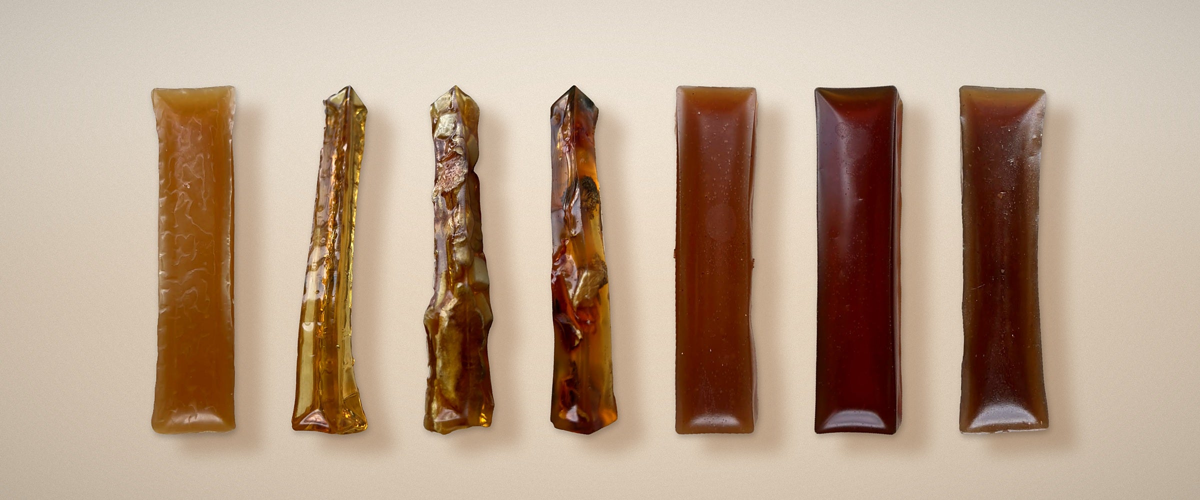 Seven bone broth hard chews for dogs standing in a row on cream, from pale golden through clear amber to deep brown. BARF INDIA