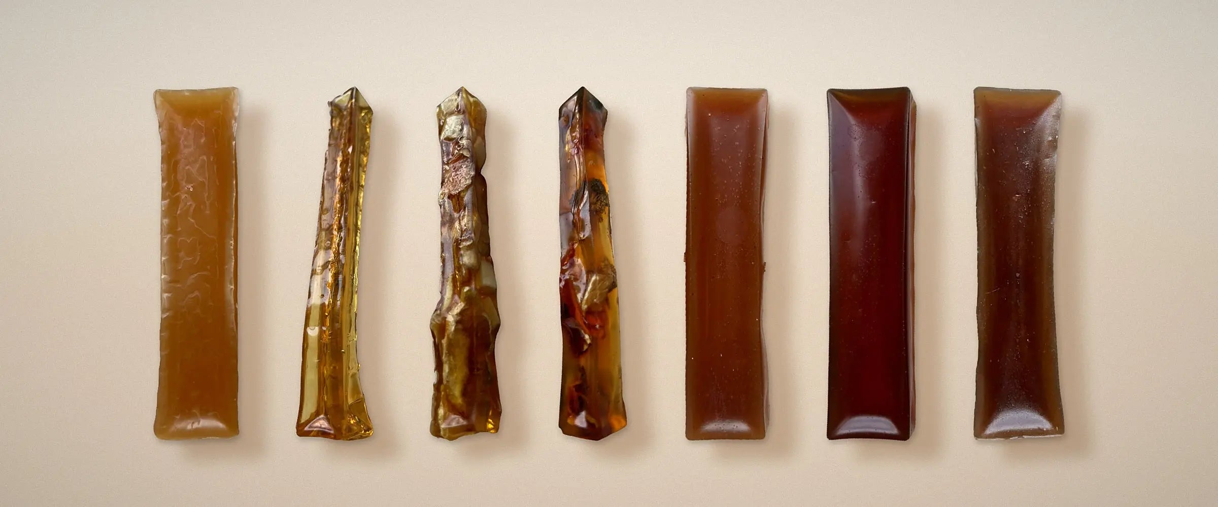 Seven bone broth hard chews for dogs standing in a row on cream, from pale golden through clear amber to deep brown. BARF INDIA