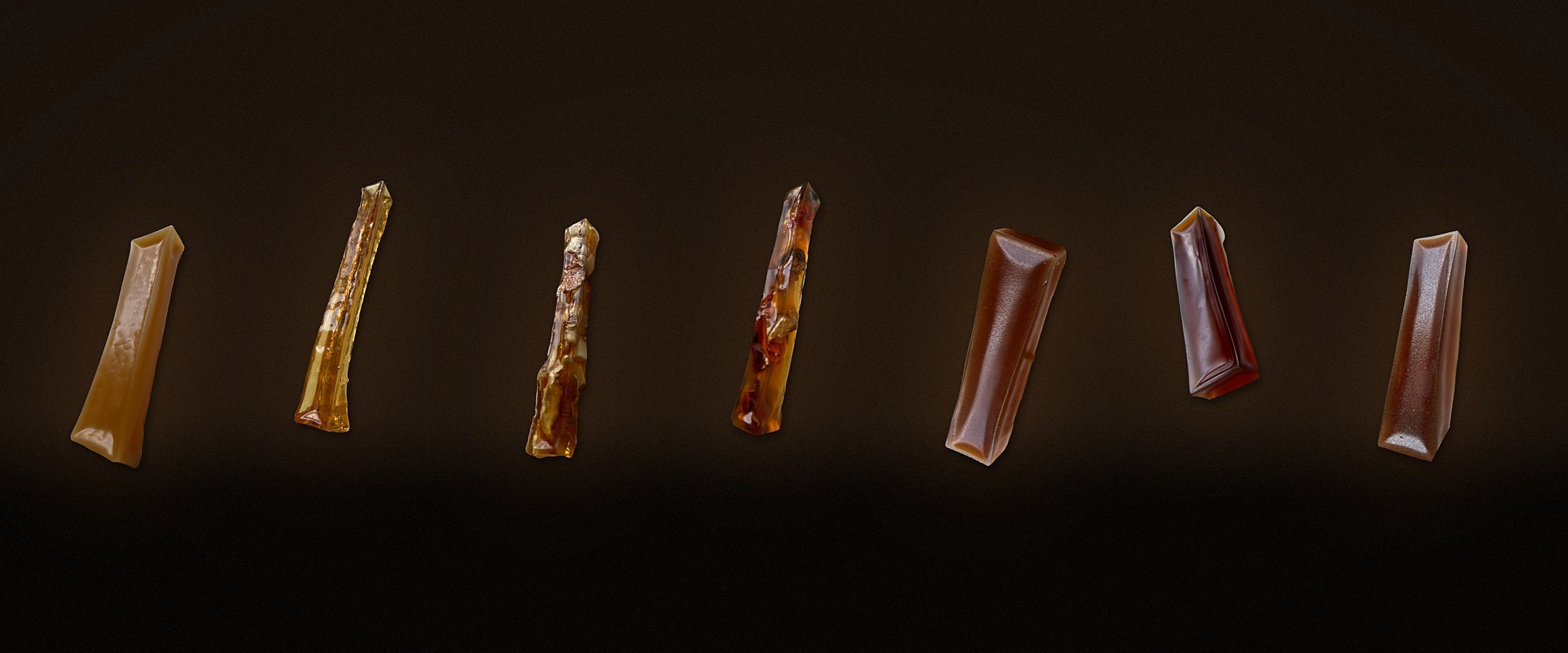 Seven bone broth dog chews standing on a dark background with a warm amber glow, including clear chews with visible pieces inside. BARF INDIA