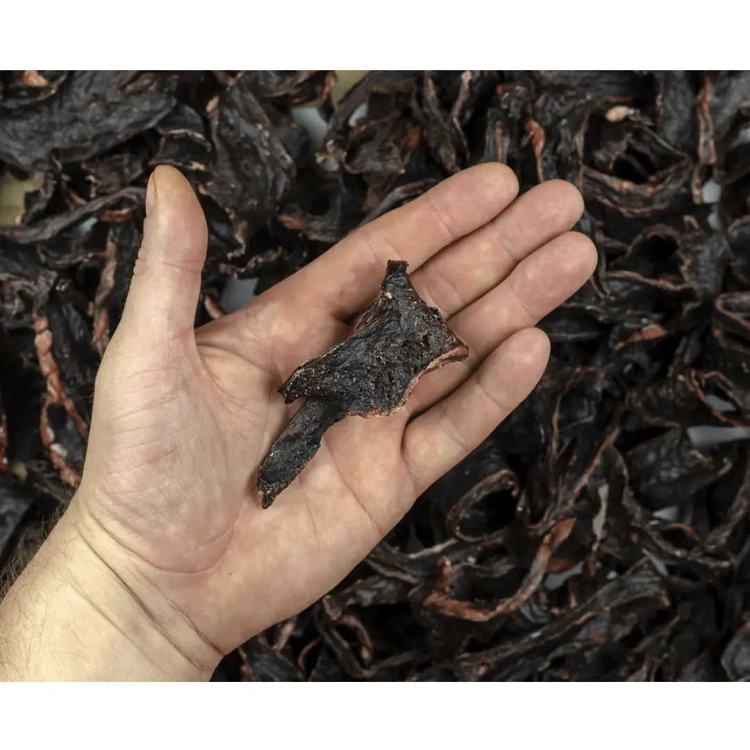 A hand holds a piece of Dog Treats Buffalo Heart - 60 gms by www.barfindia.com over a pile of similar air-dried treats—wholesome, natural snacks perfect for rewarding your dog.