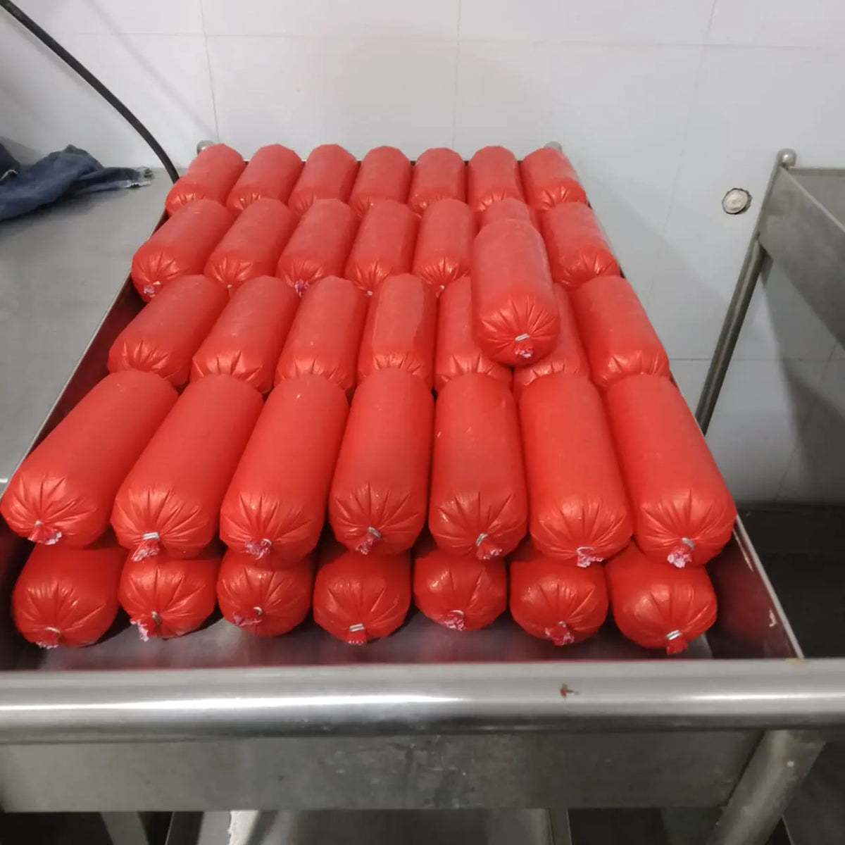 Rows of red sausage-shaped packages of Raw Dog Food - Buffalo Recipe 4 kgs from www.barfindia.com are neatly stacked on a metal table in a food processing area with white tiled walls.