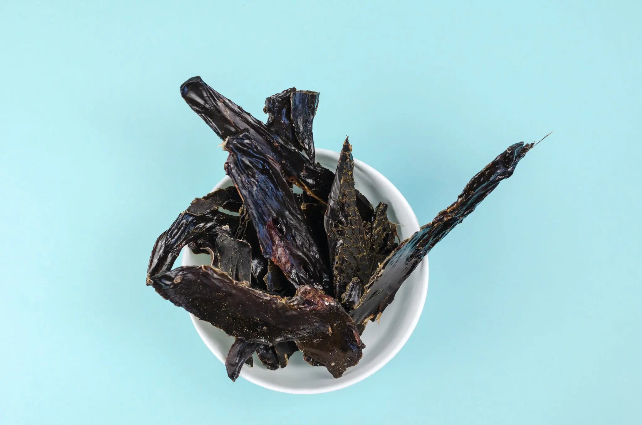A white bowl filled with dark buffalo liver jerky strips—Dog Treats - Buffalo Liver - 100 gms by www.barfindia.com—makes a protein-rich snack for dogs, captured from above on a light blue background.