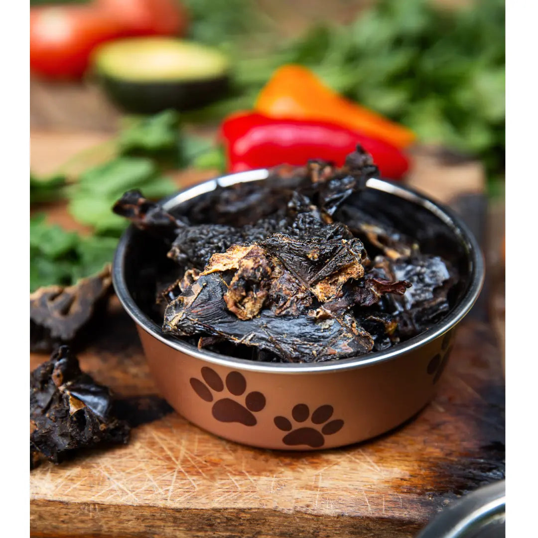 A metal pet bowl with paw prints holds www.barfindia.com Dog Treats - Buffalo Liver - 100 gms, placed on a wooden surface with blurred vegetables like red pepper and leafy greens in the background.