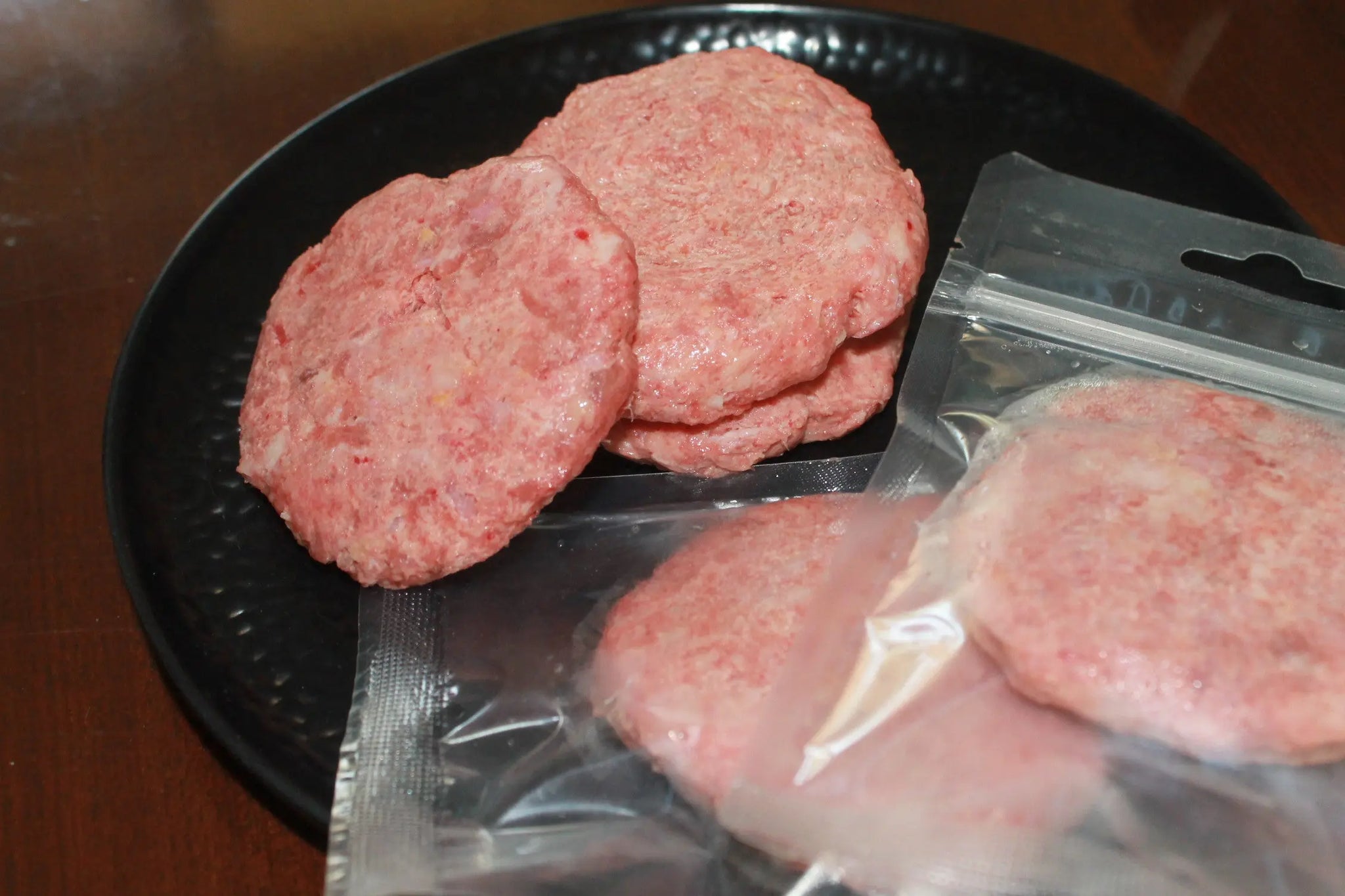 Four raw patties—two on a black plate, two vacuum-sealed—sit on wood, showcasing www.barfindia.com's Raw Cat Food - Duck and Quail, perfect for feline raw food fans.