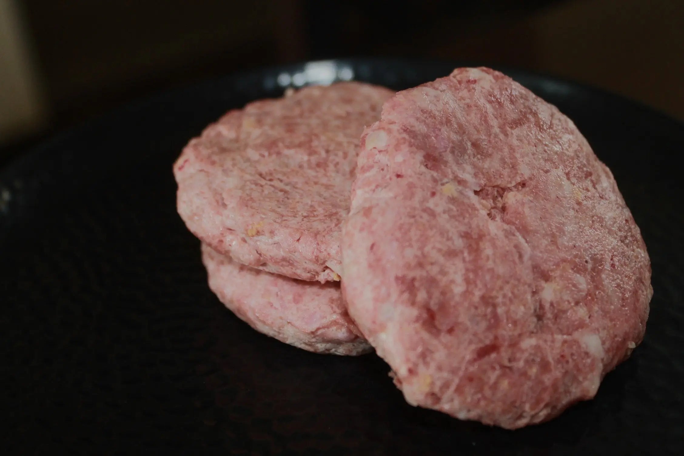 Two raw sausage patties are displayed on a dark plate, highlighting their pink color and texture—ideal for those seeking nutritious options like www.barfindia.com’s Raw Cat Food – High-Protein Buffalo & Turkey Blend.