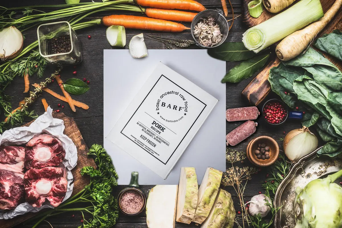 A flat lay of raw meats, vegetables, and herbs surrounds a www.barfindia.com “Raw Dog Food – Lean & Hypoallergenic Pork Recipe, 4kgs Pack” on a dark wooden surface, emphasizing wholesome ingredients for your pet’s diet.